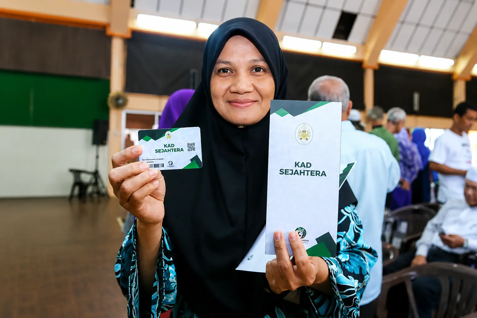 Wanita memegang Kad Sejahtera Terengganu dan risalah program rawatan kesihatan di sebuah dewan.
